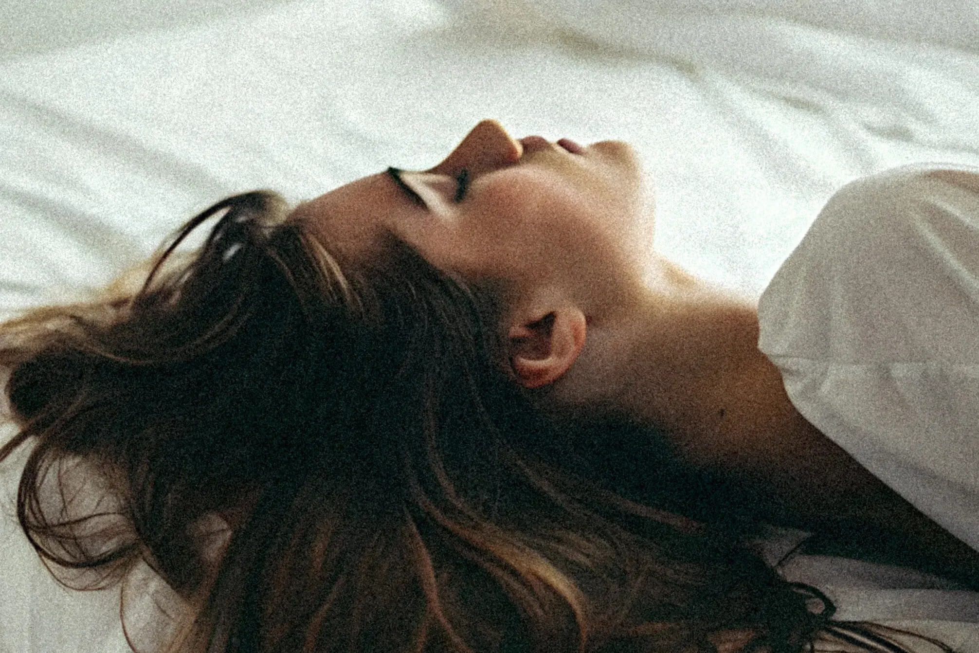 Woman lying back on white bedding with her eyes closed, deeply relaxed
